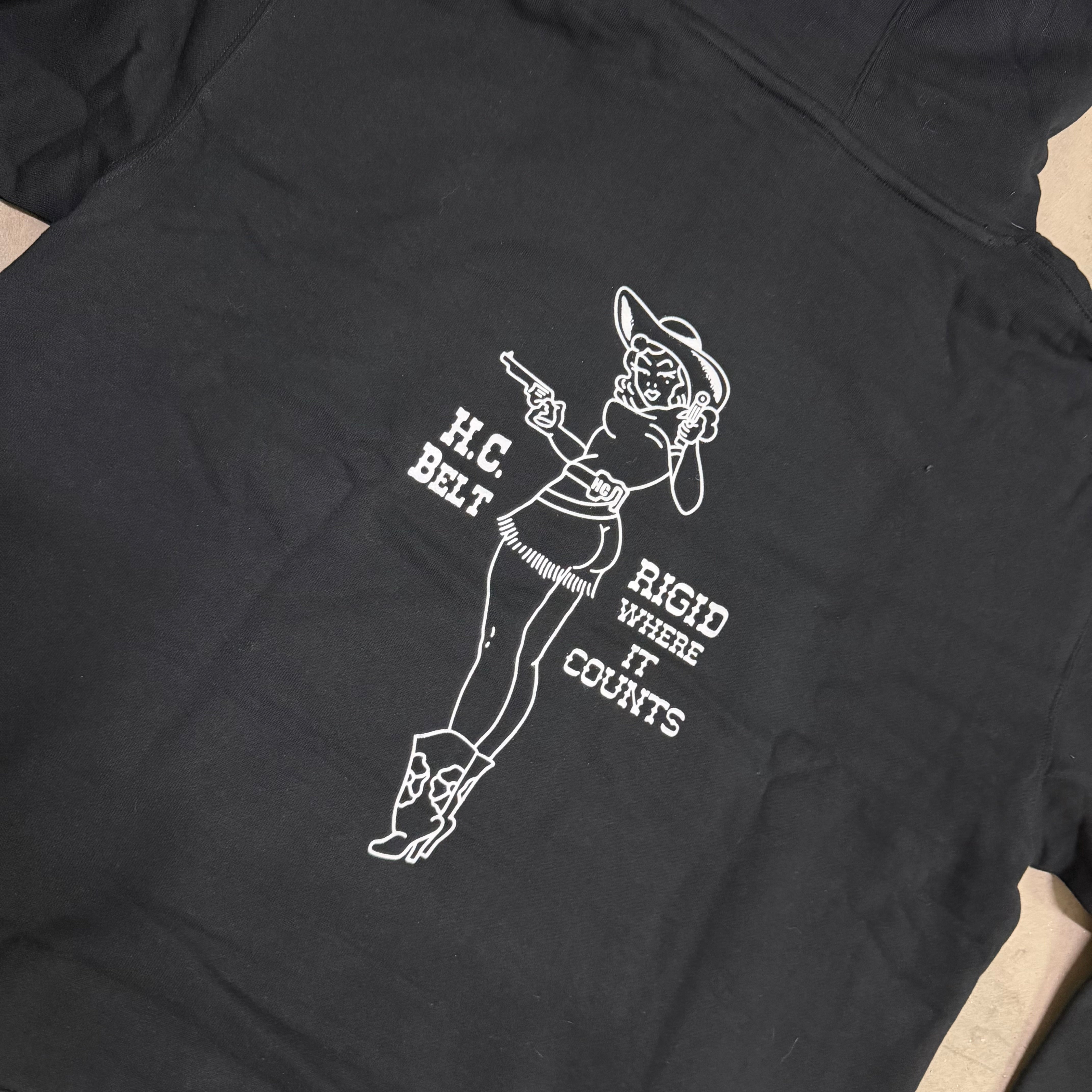 Black t-shirt with white graphic design of a woman in a cowboy outfit and text on a beige surface.